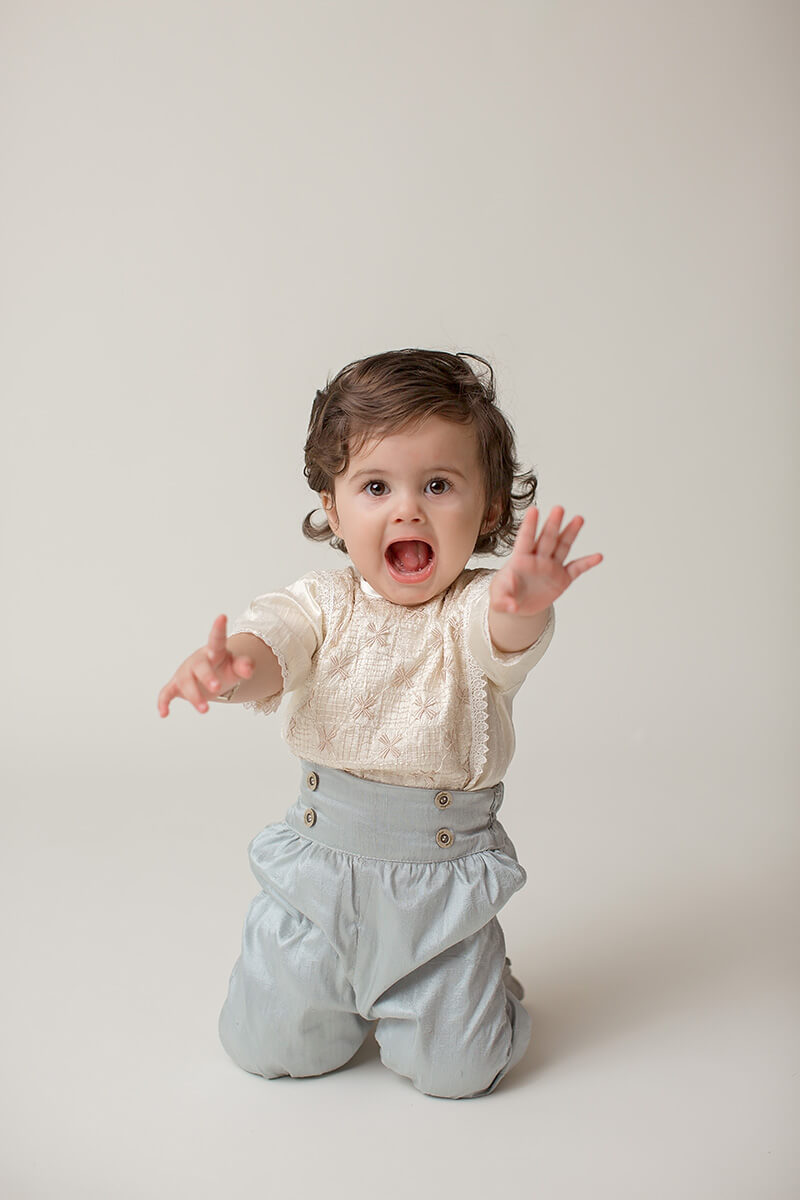 A toddler in silver pants and cream blouse plays with mouth open and arms out in a studio before visiting indoor playgrounds in Champaign, IL
