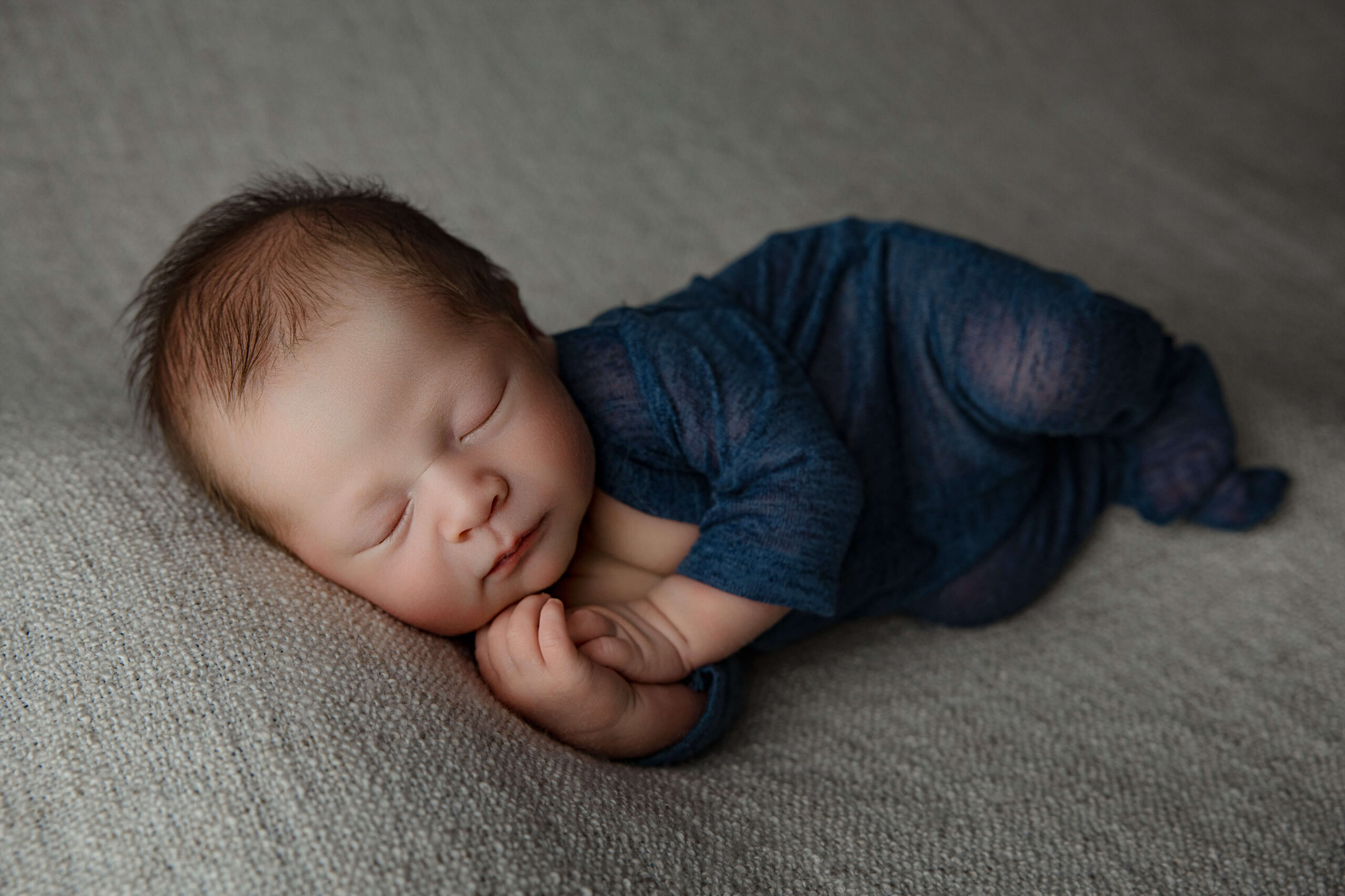 A newborn baby sleeps on his side in a blue onesie on a grey bed
