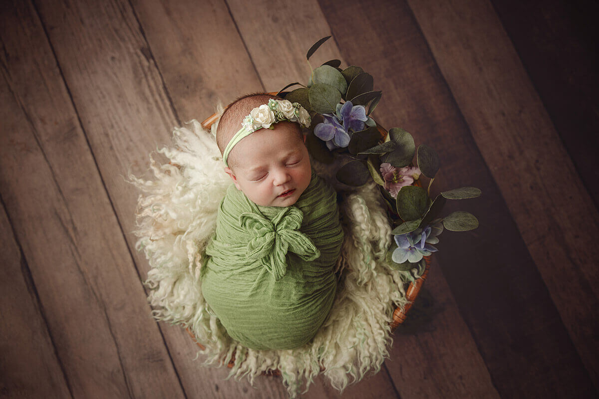 A sleeping baby girl in a green swaddle and flower headband sleeps in a wicker basket with flowers while mom enjoys pelvic floor therapy in champagin il