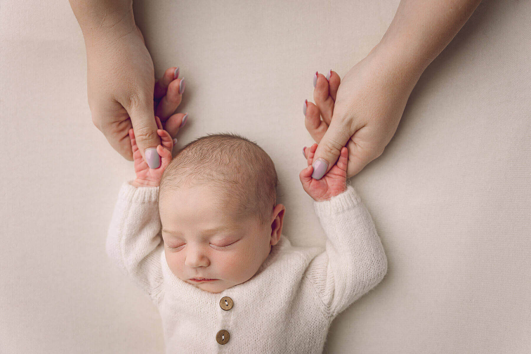 A newborn lays sleeping in a white onesie while holding mom's thumbs over his head