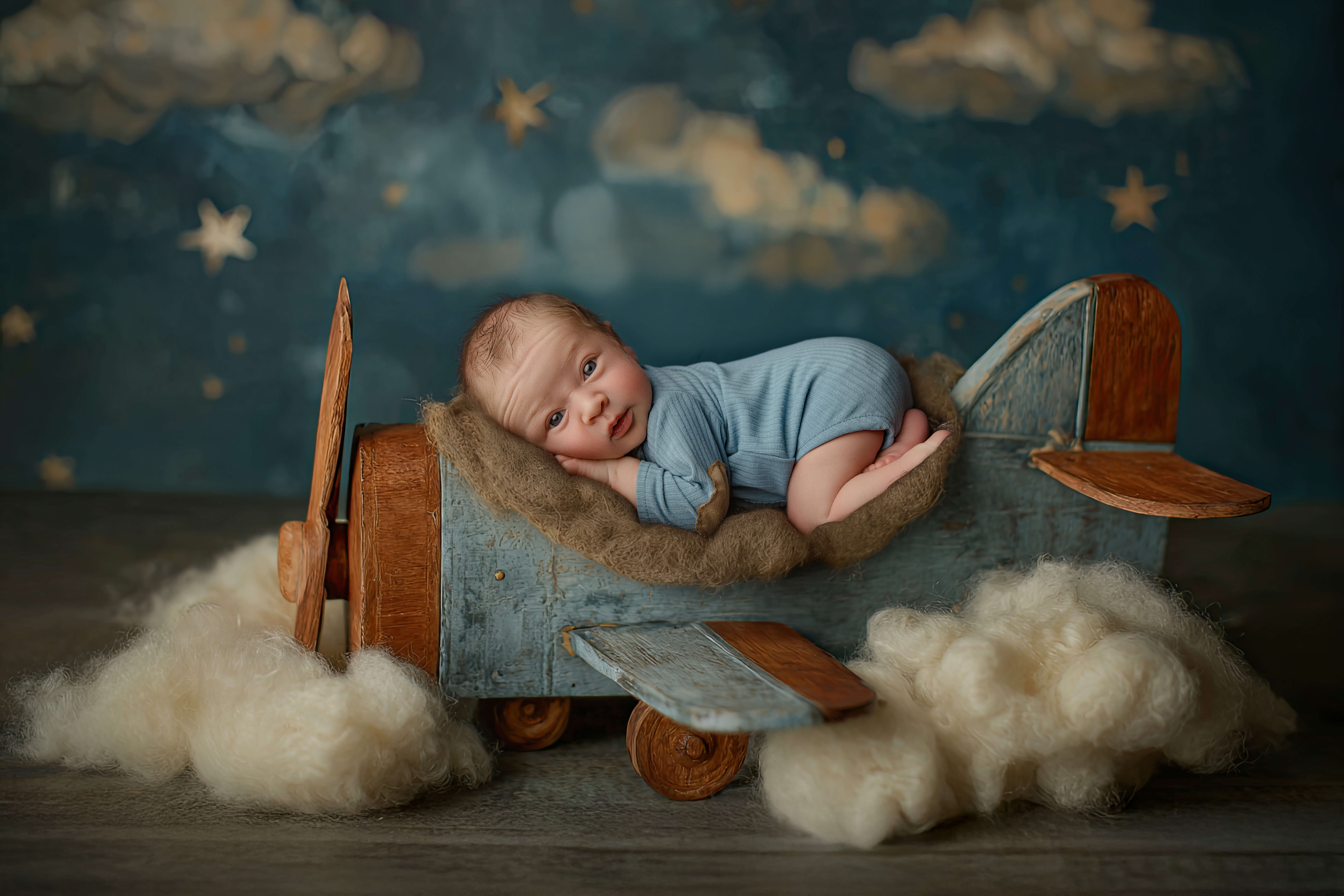 A newborn baby lays awake in froggy pose on a wooden airplane in blue pajamas