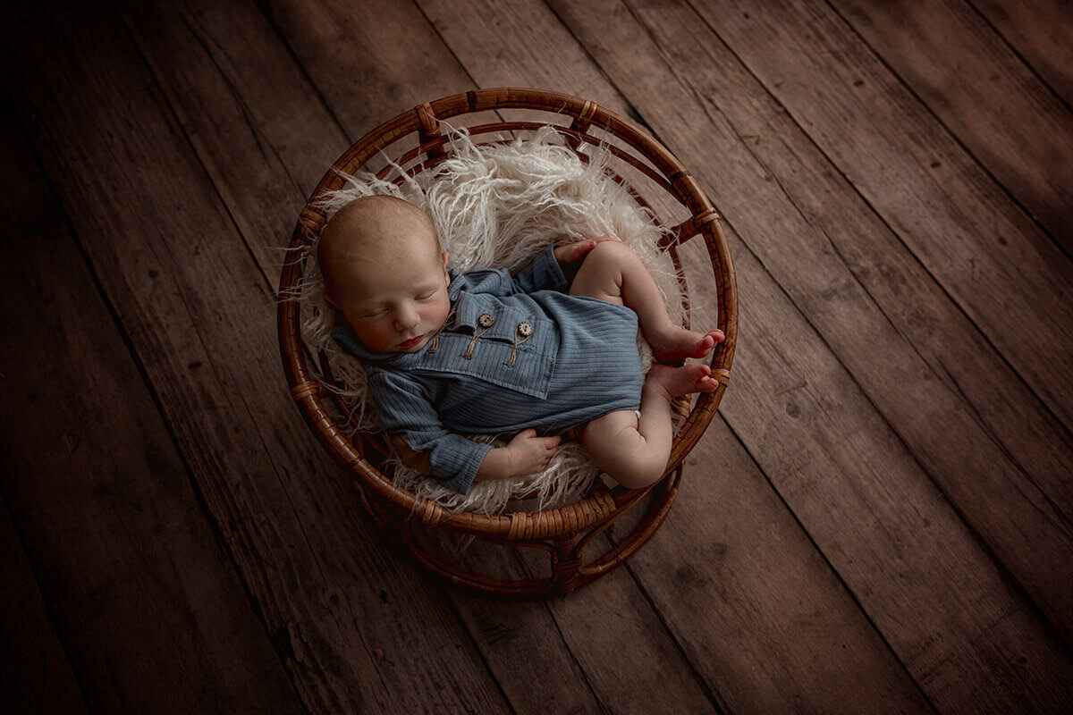 A newborn sleeps in blue pajamas in a wicker basket on wood floor thanks to prenatal chiropractor in champagin il
