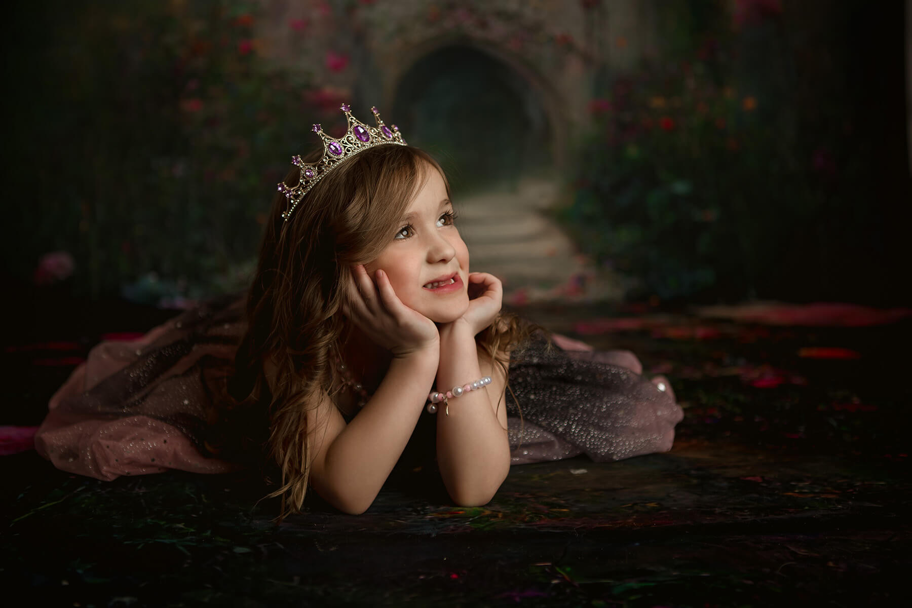 A young girl lays on the floor resting her head in her chin while wearing a long gown and purple tiara