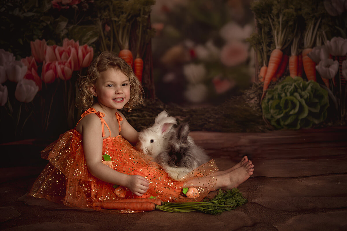 A young girl in an orange dress sits in a studio petting a furry rabbit in her lap