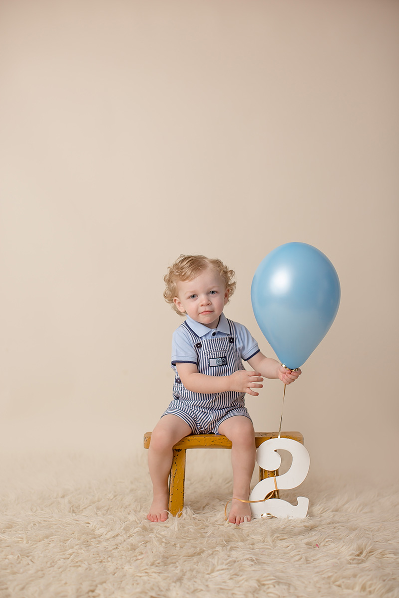 A happy toddler boy in blue overalls plays with a blue balloon in a studio while sitting on a stool