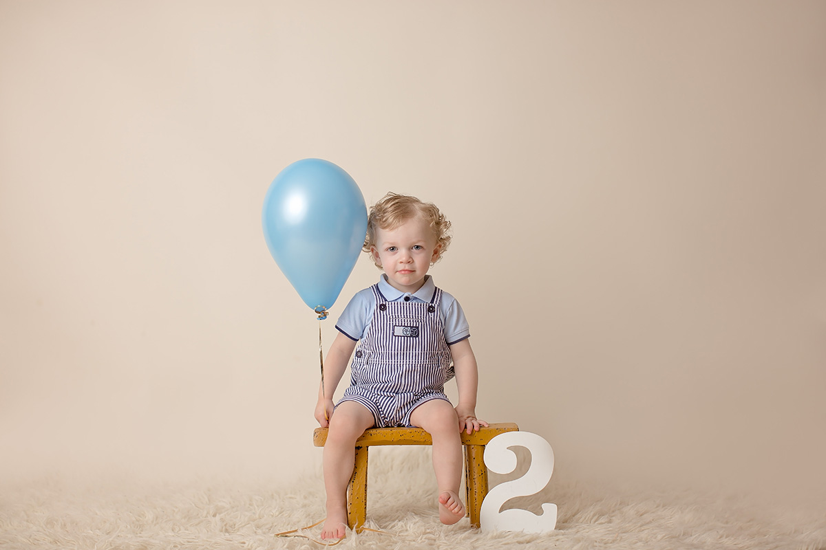 A young boy sits on a wooden stool in a studio in blue overalls and holding a balloon after enjoying a kids birthday party in Champaign, IL