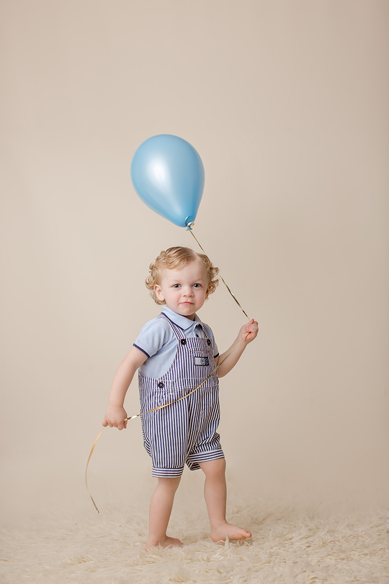 A toddler boy in blue overalls holds a blue balloon by the string while walking barefoot in a studio after his kids birthday party in Champaign, IL