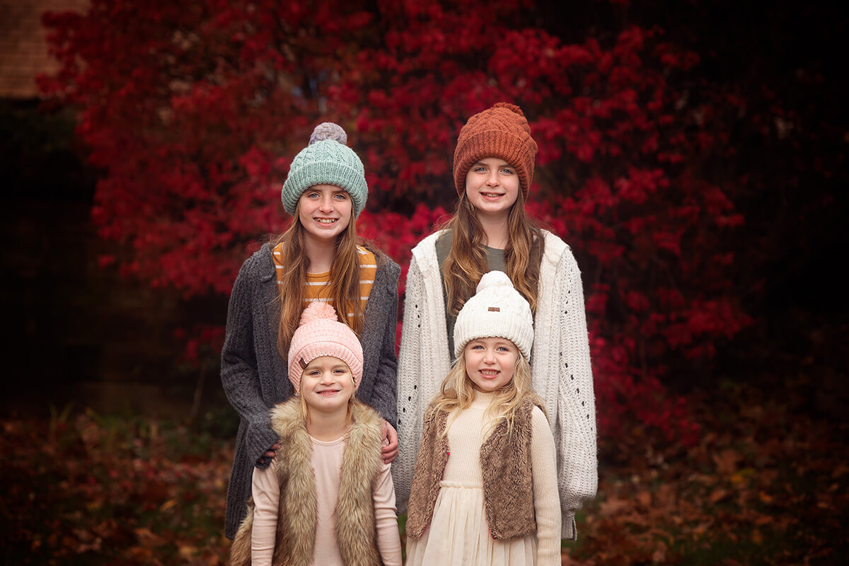Four sisters stand smiling in a lawn in front of res bush in fall wearing sweaters and beanies