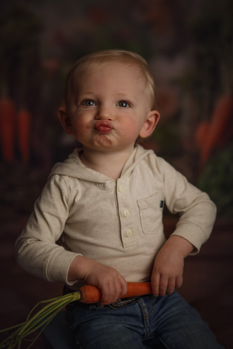 A toddler boy in a white shirt with a hood makes a silly face while sitting holding a carrot in a studio