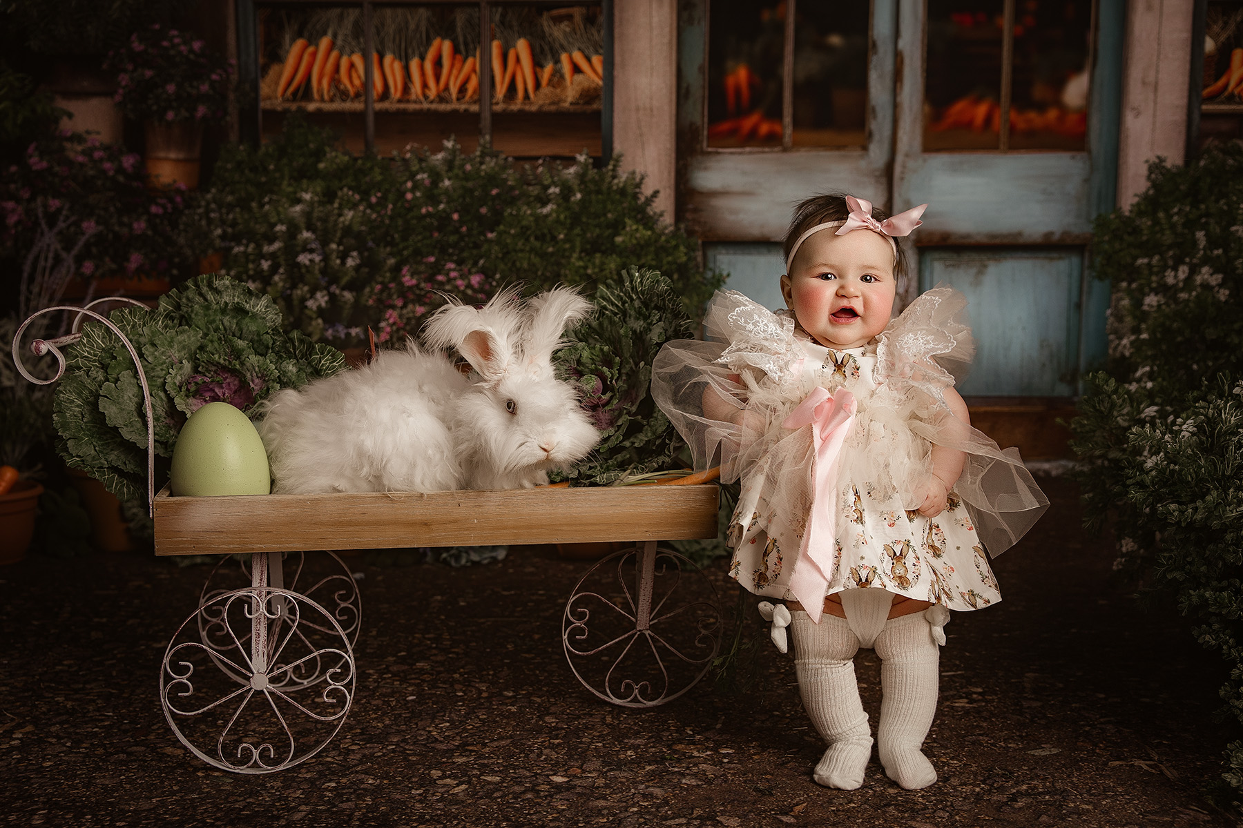 A happy baby girl in a bunny dress stands next to a fluffy bunny in a wooden wagon after exploring preschools in champaign il
