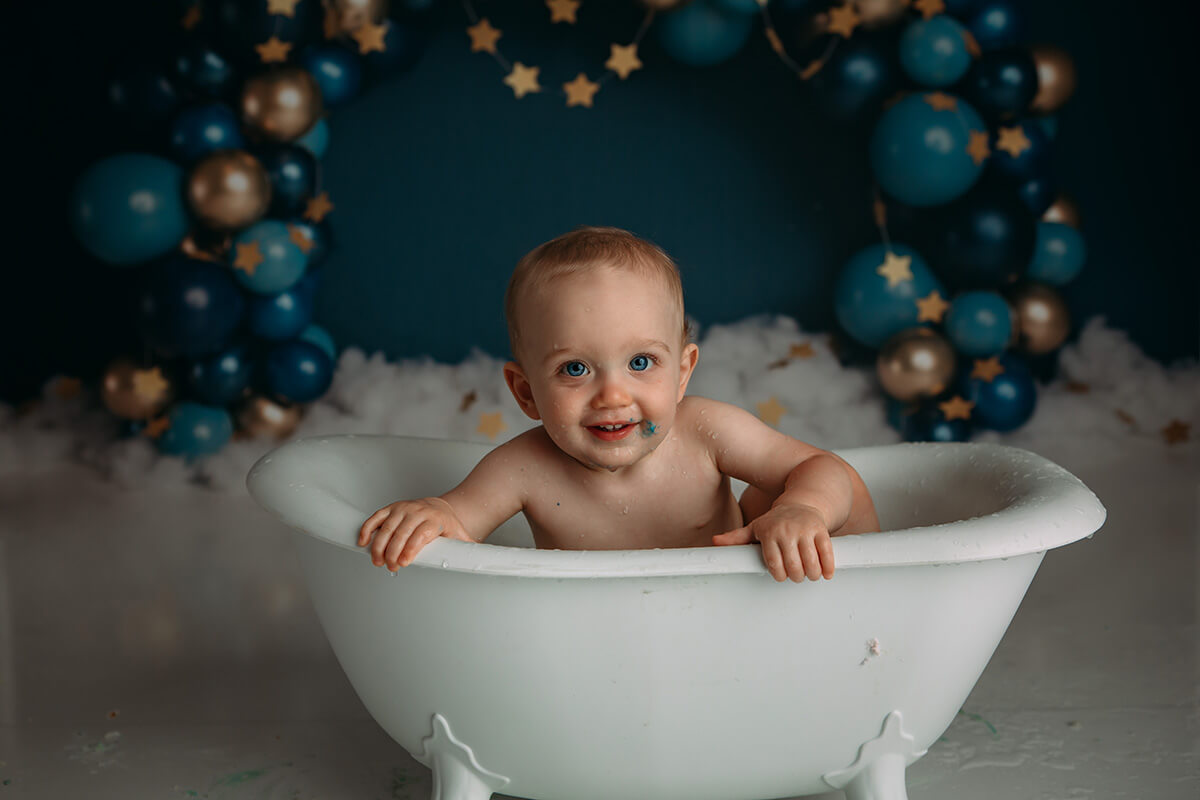 A happy toddler boy covered in blue cake icing plays in a bathtub in a studio