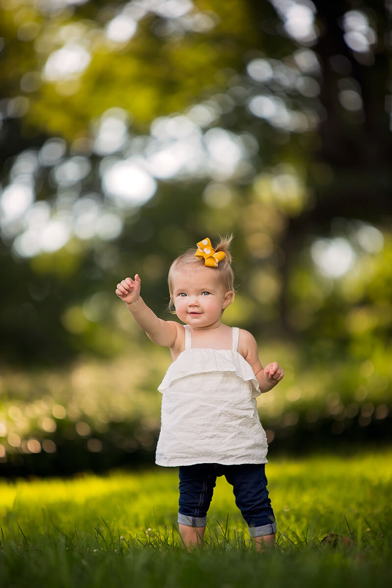 A smiling young toddler in jeans and white shirt gives a thumbs up while playing in a lawn