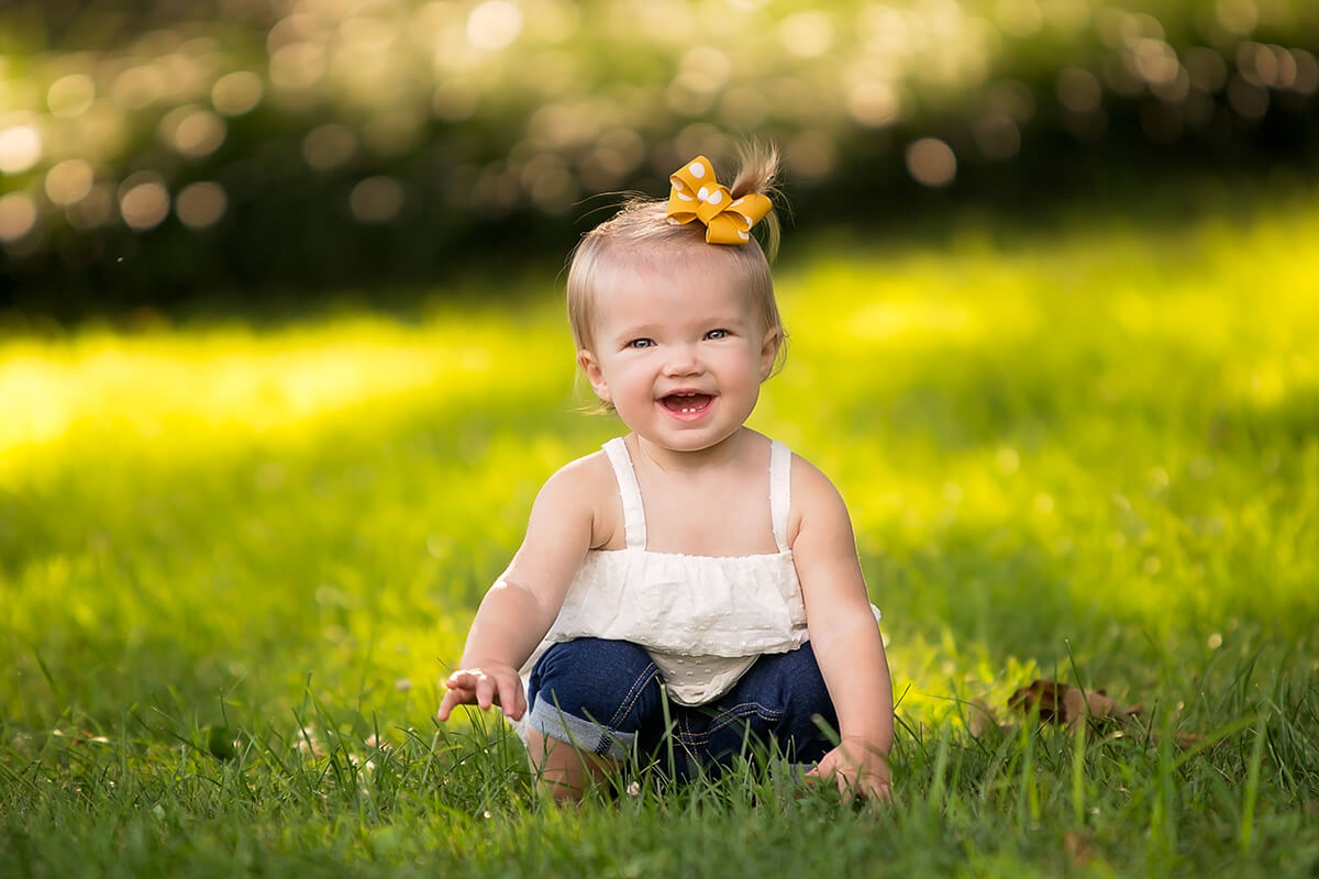 A baby girl laughs while playing in the grass in a white blouse and yellow hair bow after some swim lessons in champaign il