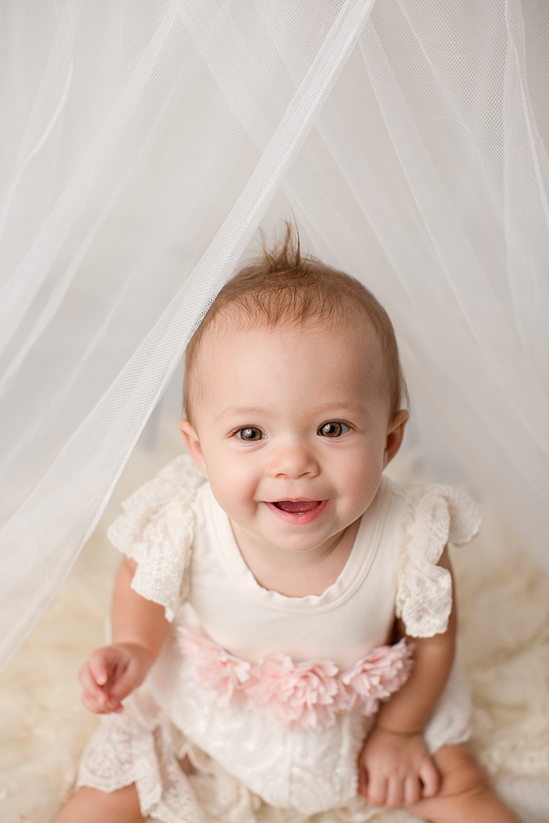 A baby girl sits in a white and pink dress under white drape smiling in a studio after visiting toy stores in Champaign, IL