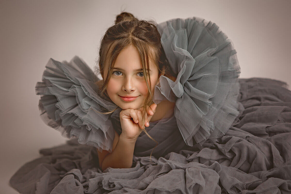 A young girl leans on her hand while laying across a studio floor in a grey tulle dress with big ruffle shoulders